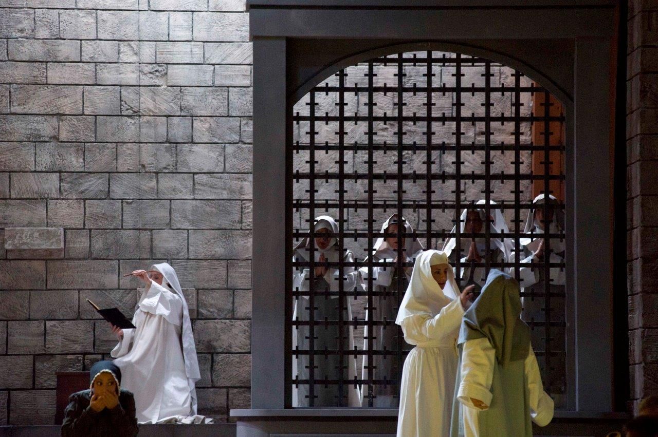 Scena di Suor Angelica: suore in abito bianco davanti a una grata di ferro, in un chiostro di pietra